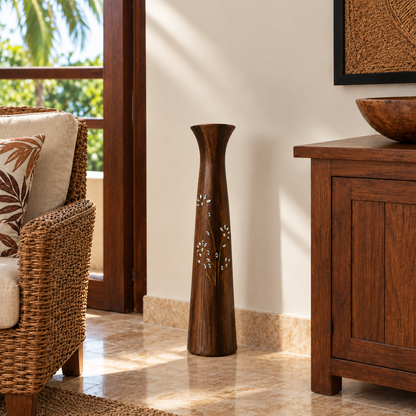 A tall, dark brown wooden decorative vase with white floral inlay, standing on a tiled floor in a warm, sunlit room next to a wicker chair and wooden cabinet.