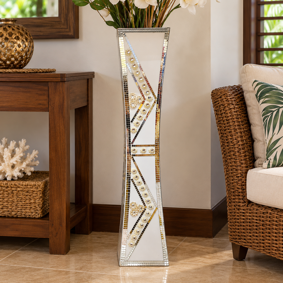 A tall, narrow floor vase with a mirrored finish and geometric gold, silver, and black mosaic accents, styled in a living room next to a wicker chair and wooden side table.