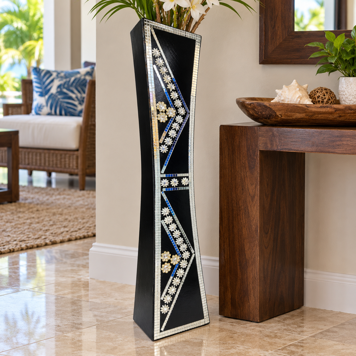 A tall, narrow black decorative vase with geometric inlay patterns in white, blue, and gold, holding white flowers, set in a modern living room next to a wooden console table.