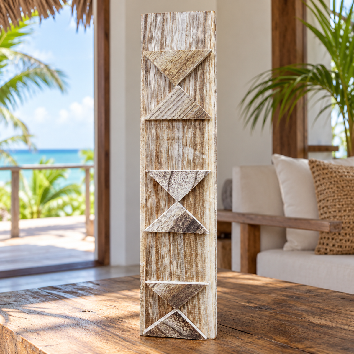 A tall, narrow decorative art piece with carved geometric patterns, standing on a rustic wooden table in a bright, modern outdoor living space with ocean views in the background.