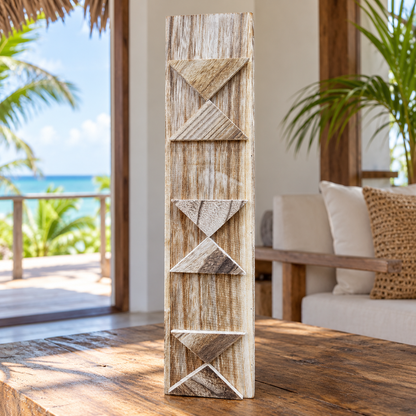 A tall, narrow decorative art piece with carved geometric patterns, standing on a rustic wooden table in a bright, modern outdoor living space with ocean views in the background.