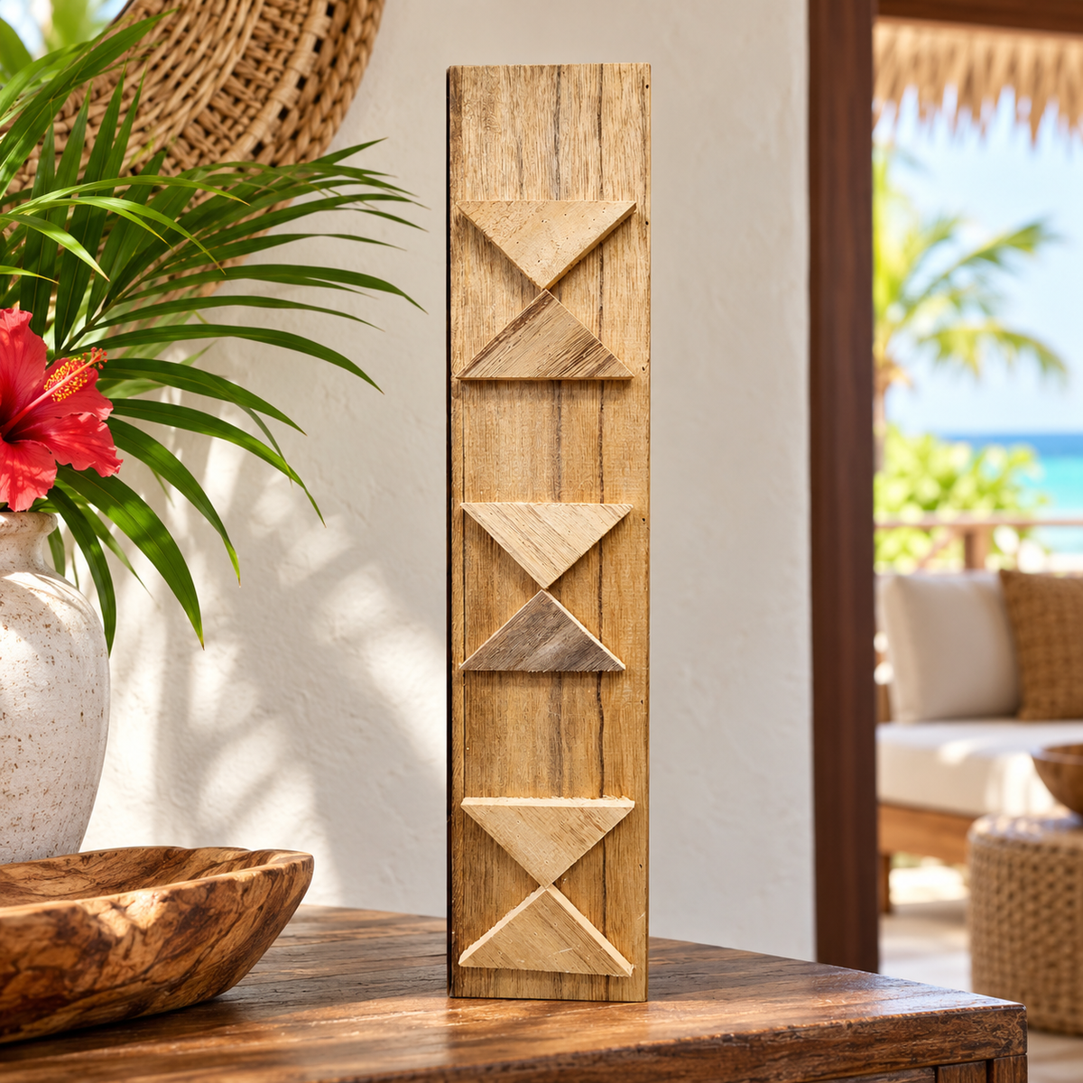 A tall rectangular decorative wall piece with carved geometric patterns, displayed on a table in a tropical patio setting with a plant and ocean view in the background.