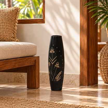 A tall, slender black ceramic vase with gold botanical and star patterns, standing on a polished stone floor in a sunlit, modern interior setting.